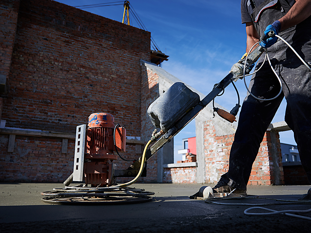 Cropped picture of man laborer in work overalls using power trowel machine while screeding floor in new building. Male worker finishing concrete surface with troweling machine at construction site.